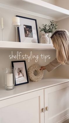 a woman standing on top of a white shelf next to a vase and framed pictures