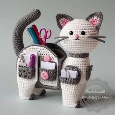 a crocheted white cat with pink ears and tail holding a pen holder filled with office supplies