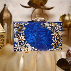 Overview Make a stunning statement on your special day with this luxurious blue acrylic bridal clutch. Crafted with exquisite detail, this minaudiere features a rich blue hue complemented by intricate gold floral accents and dazzling crystal embellishments. The structured design and elegant gold clasp closure make it a chic and practical accessory for brides, bridesmaids, or any evening event. Each clutch is carefully handcrafted, showcasing a combination of modern resin artistry with timeless g Glamorous Blue Evening Bag For Gift, Luxury Blue Rectangular Clutch, Glamorous Blue Rectangular Clutch, Formal Handmade Blue Clutch, Handmade Blue Evening Bag For Formal Occasions, Handmade Blue Clutch For Formal Occasions, Handmade Blue Clutch For Formal Events, Blue Glamorous Party Clutch, Blue Luxury Evening Bag As Gift