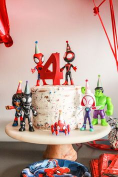 a birthday cake with spiderman figurines on it and balloons in the background