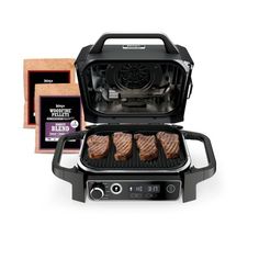 an electric grill with four steaks cooking on it and a box of meat next to it