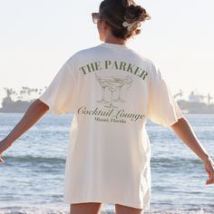 a woman standing on the beach with her back to the camera, wearing a t - shirt that says the parker cocktail lounge