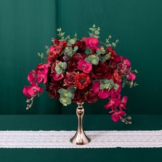 a gold vase filled with red flowers on top of a white table cloth next to a green wall