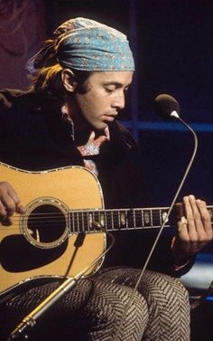 a man sitting in front of a microphone while holding a guitar and wearing a bandana