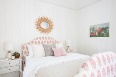 a bedroom with white walls, pink and white bedding, and a sun mirror on the wall