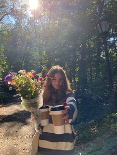 a woman holding a cup and flowers in her hands while standing on a path surrounded by trees