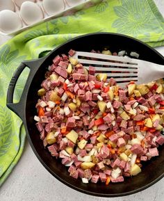 a skillet filled with ham and vegetables on top of a green towel next to eggs