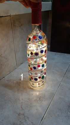 a hand holding a bottle that has been decorated with beads and lights on it, sitting on a tile floor