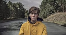 a young man in a yellow jacket standing on the side of a road with trees behind him