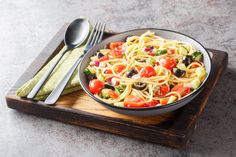 a bowl of pasta with tomatoes, olives and asparagus on a wooden tray