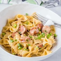 a white bowl filled with pasta covered in sausage and peas next to a fork on top of a table