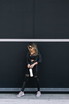 a woman standing in front of a black wall wearing leggings and holding a bottle