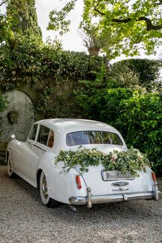 an old white car with flowers on the hood parked in front of a stone wall