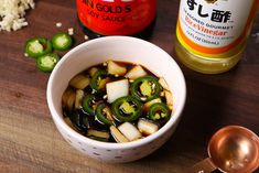 a white bowl filled with vegetables next to a bottle of hot sauce and spoon on a wooden table