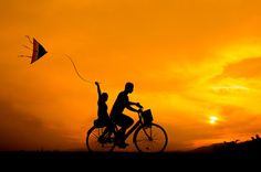 two people riding bikes with a kite flying in the sky behind them at sunset or dawn