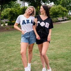 two young women standing next to each other in the grass with trees and buildings behind them