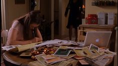 a woman sitting at a table covered in papers and magazines with her head on her hand