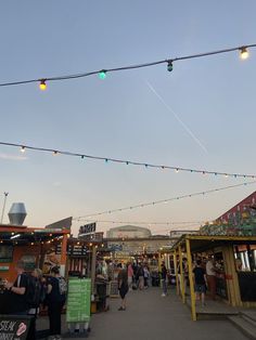 people are walking around an outdoor market with lights strung over it and buildings in the background