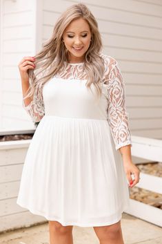 a woman wearing a white dress with sheer lace sleeves and heels standing on the sidewalk