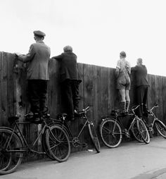 several men standing on bicycles in front of a wooden fence and looking at the sky