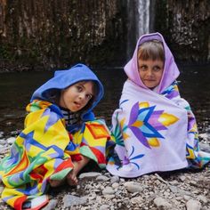 two young children sitting on the rocks under a waterfall wrapped in colorful blankets and smiling at the camera