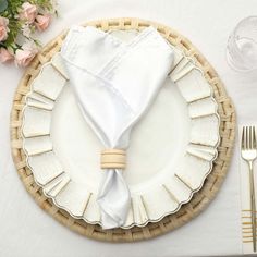 a place setting with white napkins and silverware on a wicker plate, along with pink flowers