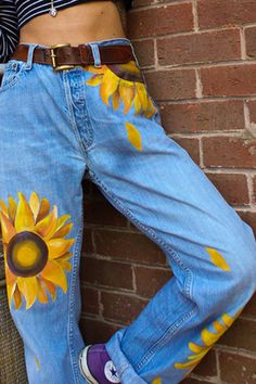 a woman leaning against a brick wall with sunflowers painted on her jeans