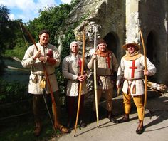 three men dressed in period clothing holding spears and arrows, standing next to a stone building