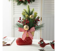 a vase filled with pine branches and berries on top of a table next to a bird figurine