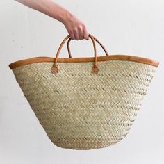 a hand holding a woven basket with leather handles on the handle is shown in front of a white wall