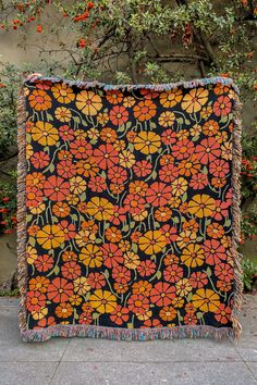 an orange and black blanket with flowers on it sitting in front of a tree filled with leaves