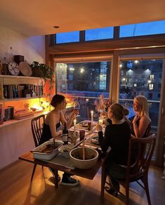 three women sitting at a table with food and drinks in front of them, looking out the window