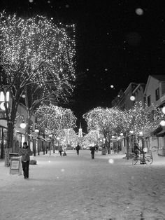 people walking down a snowy street at night with lights on trees and buildings in the background