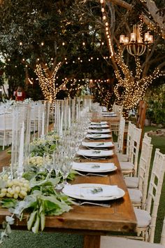 a long wooden table topped with lots of white plates and place settings covered in greenery