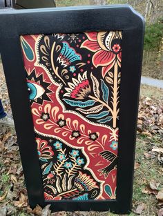 an artisticly painted wooden box sitting on the ground in front of some leaves and trees