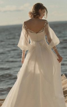 a woman in a white wedding dress standing on the beach looking at the water with her back to the camera