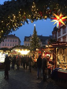 people are walking through an outdoor market at christmas time with lights on trees and buildings in the background