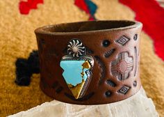 A one of a kind 1.5 inch hand stamped and stained leather cuff in a dark brown, with a mixed southwestern stamping pattern.  The cuff has an artisan handmade sterling silver piece that has a stamped symbol of the sun on the top and an arrow  with a beautiful piece of Baja turquoise in the center that measures 1.25 inches in length and .75 at the widest point.  All of our bracelets are hand crafted by us from the finest leather and constructed to last.  We take our time researching pieces for our Western Style Brown Cuff Bracelet For Festivals, Artisan Brown Cuff Bracelet With Concho, Artisan Brown Concho Cuff Bracelet, Western Style Brown Cuff Bracelet As Gift, Rustic Brown Cuff Bracelet With Concho, Southwestern Hand Tooled Brown Cuff Bracelet, Rustic Stamped Leather Bracelet, Bohemian Stamped Brown Cuff Bracelet, Brown Bohemian Stamped Cuff Bracelet