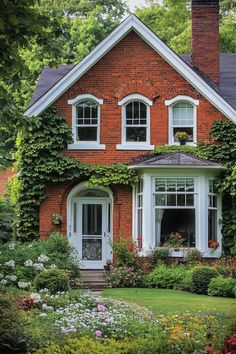 a red brick house with white windows surrounded by flowers and greenery on the front lawn