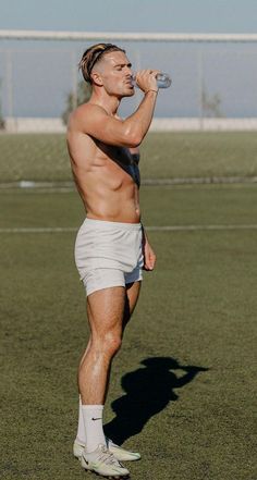 a shirtless man drinking from a water bottle on a field with grass and trees in the background