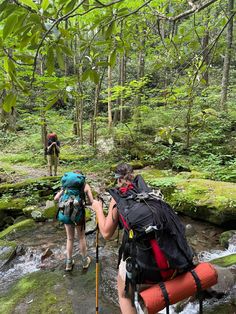 two people with backpacks cross a small stream in the woods while another person stands behind them