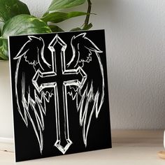 an artistic cross with wings on a black background art boarder next to a potted plant