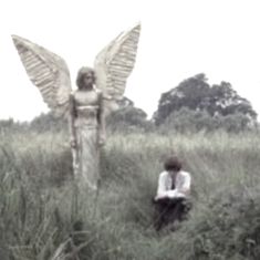 an angel statue sitting in the middle of a field next to a woman with her arms crossed