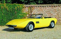 a yellow sports car is parked in front of a brick wall and shrubbery behind it