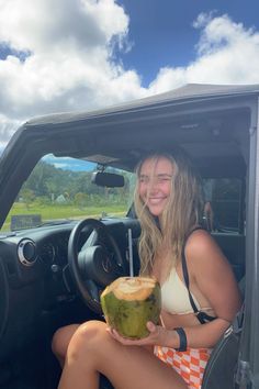 a woman sitting in the driver's seat of a car holding a coconut and a drink