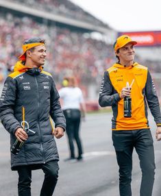 two men in yellow and black jackets walking on a race track with one holding a drink