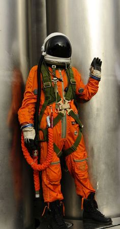 an orange astronaut suit standing in front of a metal tank with his hand on the side