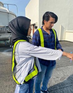 two people in safety vests are standing on the pavement and pointing at something with one hand