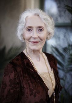 an older woman with white hair wearing a brown dress and pearls smiles at the camera