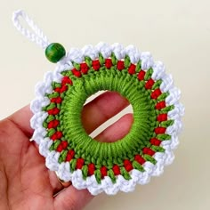 a hand holding a green and red crocheted christmas wreath ornament on a white background